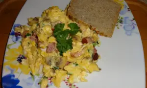 Pyszna jajecznica z pieczarkami, boczkiem i szczypiorkiem, udekorowana natką pietruszki, podana z kromką chleba.