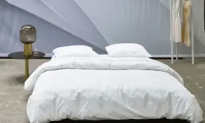 Ein Bett mit weißer Perkal-Bettwäsche, gesteppt mit feinen Linien. Daneben ein Nachttisch mit Lampe und Parfüm.