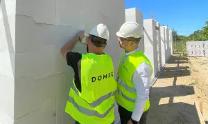 Dwóch mężczyzn w kamizelkach odblaskowych na placu budowy sprawdza postęp prac przy ścianie z białych pustaków.