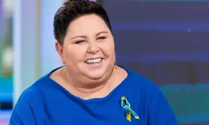 Uśmiechnięta Dorota Wellman w niebieskim swetrze z przypinką w barwach Ukrainy. Jej wykształcenie i doświadczenie pomagają jej w pracy.