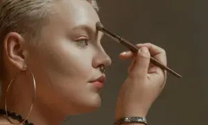 Kobieta z krótkimi blond włosami i kolczykiem w nosie nakłada bronzer na czoło, ucząc się, jak wyszczuplić twarz bronzerem.