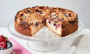 Ein köstlicher Beerenkuchen vom Blech, bestreut mit Mandelsplittern, steht auf einem Tortenständer. Daneben eine Schale mit frischen Beeren.