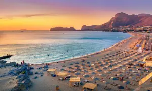 Traumhafter Badeurlaub im Mai: Sonnenschirme säumen den Sandstrand, das Meer glitzert, und die Berge im Hintergrund tauchen in warmes Abendlicht.