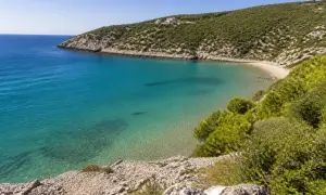 Vista dall'alto della spiaggia di Procinisco, con acque cristalline turchesi che lambiscono una sabbia dorata, incorniciata da colline verdi e rocciose.