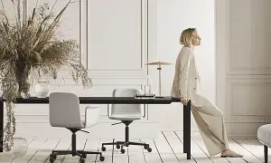 Eine Frau in einem beigen Anzug sitzt auf einem schwarzen Tisch. Der Raum ist in neutralen Farben gehalten und mit Trockenblumen und Glasvasen dekoriert.