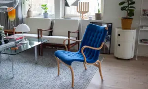 En blå Bruno Mathsson stol med böjda träarmstöd står på en grå matta. Bredvid syns vita soffor och ett glasbord.