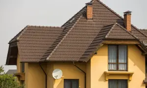Dach naczółkowy konstrukcja z brązowej dachówki, z ceglanym kominem i anteną satelitarną na żółtej ścianie domu.
