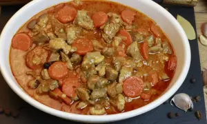 Pyszny gulasz z karkówki z marchewką w aromatycznym sosie, podany w białej misie. Obok czosnek, liść laurowy i ziele angielskie.