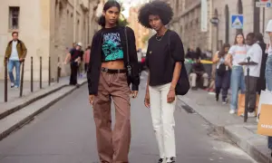 Zwei Frauen in lässiger Kleidung, eine mit brauner Cargohose und Crop-Top, die andere mit weißer Hose und schwarzem Shirt, stehen auf einer Straße.