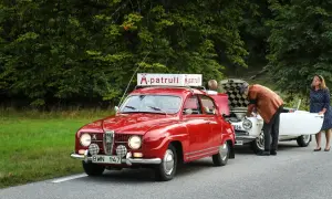 Röd Saab 96 V4 med "M-patrull" skylt på taket. En man tittar i motorhuven på en vit bil, en kvinna står bredvid.