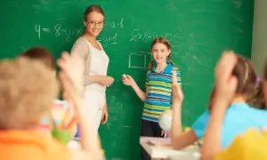 Nauczycielka dyplomowana z uśmiechem obserwuje uczennicę przy tablicy. To przykładowa ocena pracy nauczyciela.