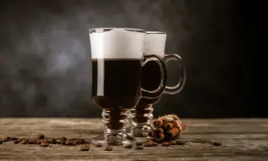Två glas med irish coffee, toppade med grädde, står på ett träbord. Kaffebönor och kanelstänger ligger utspridda.