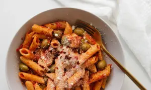 Makaron penne w sosie pomidorowym z oliwkami i parmezanem, podany w misce ze złotym widelcem.