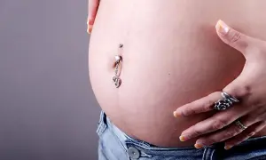 Brzuszek w ciąży z kolczykiem w pępku, ozdobionym serduszkiem. Dłoń z pierścionkami delikatnie dotyka skóry.