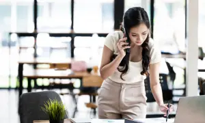 Kobieta rozmawia przez telefon, pochylona nad biurkiem z laptopem i dokumentami. Wygląda na zmartwioną, jakby nie mogła wysłać sms do jednej osoby.