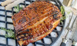 Perfekt gegrillter Schweinebauch mit knuspriger Schwarte liegt auf einem Grillrost. Kräuter und Grillbesteck deuten auf ein köstliches Mahl hin.