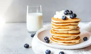 Ein Stapel goldbrauner Protein Pfannkuchen, garniert mit Sahne und Blaubeeren, daneben ein Glas Milch.