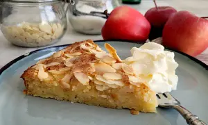 Ein Stück Apfelkuchen mit mandelkruste, garniert mit Sahne, auf einem blauen Teller. Im Hintergrund Äpfel und Mandelblättchen.