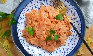 Farfalle al salmone cremose, guarnite con prezzemolo fresco, servite su un piatto blu e bianco con una forchetta dorata.