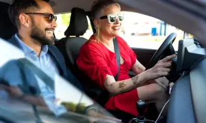 Kobieta w czerwonej sukience i okularach prowadzi auto, obok siedzi mężczyzna w okularach. Qarson w tle.
