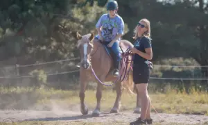Dziewczynka na koniu podczas jazdy na lonży, instruktorka z biczem i lonżą obok.