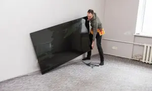 Mężczyzna w pasie narzędziowym trzyma duży, czarny telewizor przy ścianie.