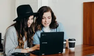 Två unga kvinnor diskuterar sin skuldkvot och bolån framför en laptop, med kaffekoppar på bordet.