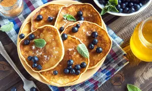 En hög med nyttiga pannkakor toppade med blåbär och pudersocker. Bredvid står en burk honung och en skål med mer blåbär.