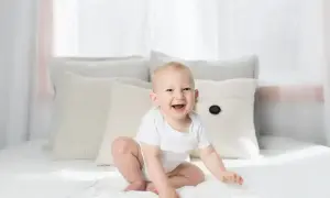 Uśmiechnięte dziecko w białym body siedzi na łóżku. To uroczy widok, który może inspirować, jak pomóc dziecku w nauce siadania.