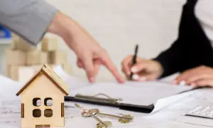 Un perito per valutazione casa indica un documento mentre un'altra persona firma. Chiavi e modellino di casa sul tavolo.