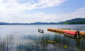 Ein Steg mit einem roten Boot am Laacher See. Schilf ragt aus dem ruhigen Wasser, das die bewaldeten Hügel am Horizont spiegelt.