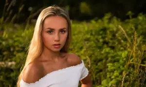 Młoda kobieta z długimi blond włosami, ubrana w białą, odkrywającą ramiona bluzkę, patrzy prosto w obiektyw na tle zielonej, rozmytej roślinności.