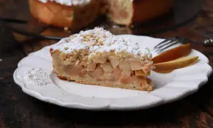 Ein Stück apfelkuchen sehr fein, mit Streuseln und Puderzucker bestäubt, auf einem weißen Teller mit Gabel und Apfelscheiben.