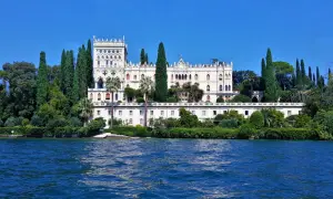 Villa neogotica su un'isola del Lago di Garda, circondata da cipressi e vegetazione lussureggiante, con il blu intenso dell'acqua in primo piano.