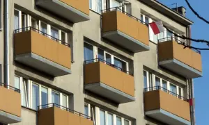 Szare bloki z brązowymi balkonami, typowe dla mieszkania komunalnego. Widać polską flagę.
