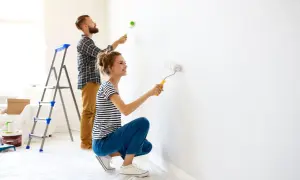 Para malująca białą ścianę wałkiem, mężczyzna na drabinie, kobieta kuca.