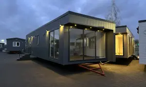 Nowoczesny dom z kontenerów, z dużymi przeszkleniami, oświetlony wieczorem. Obok stoją inne modułowe budynki.