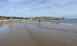 Persone passeggiano sulla battigia di Cala Molinella, con un trabucco sullo sfondo.