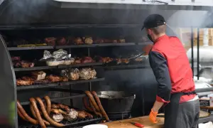 Mężczyzna w masce i rękawiczkach obsługuje wędzarnię z metalowej szafki, pełną mięs i kiełbas.