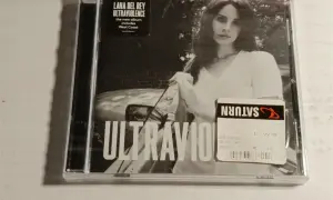 Okładka płyty Lana Del Rey Ultraviolence. Na czarno-białym zdjęciu artystka w białej koszulce, obok naklejka Saturn z ceną.