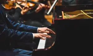Pianista w eleganckim garniturze gra na fortepianie, a przed nim leżą nuty do nauki gry na pianinie. W tle orkiestra smyczkowa.