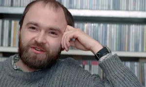 Tomasz Beksiński, zamyślony, z ręką opartą o czoło, na tle regałów z płytami. Jego spojrzenie zdaje się wybiegać poza kadr, jakby rozmyślał o przemijaniu i śmierci.