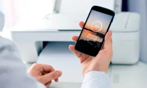 Dłoń trzyma smartfon z napisem "Sending to printer", obok drukarka drukuje dokument. Tak łatwo jest drukować z telefonu.