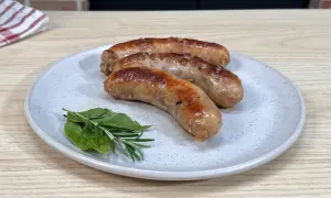 Tre salsicce dorate e succulente, appena cotte in padella, riposano su un piatto con rosmarino e alloro.
