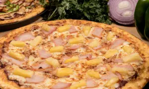 Pizza hawajska składniki: kawałki ananasa i szynki na serze. W tle inna pizza i warzywa.