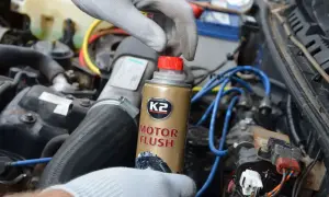 Ręka w rękawiczce trzyma puszkę płukanki do silnika K2 Motor Flush na tle silnika samochodowego.