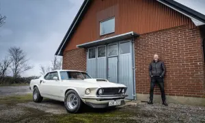 En man står stolt bredvid sin vita Ford Mustang Boss 429 framför en röd tegelbyggnad.