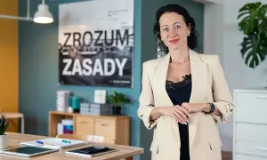 Kobieta w beżowej marynarce, obok niej na biurku leży tablet i notatnik, który może służyć jako księga przychodów i rozchodów.