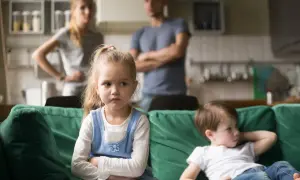 Zazdrość u dzieci w wieku szkolnym. Dziewczynka z założonymi rękami patrzy w bok, obok siedzi chłopiec. Rodzice w tle.