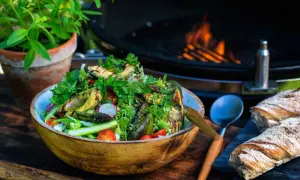 En härlig somrig sallad med grillade grönsaker, tomater och rädisor i en träskål. Bredvid ligger nybakat bröd och en grill med flammor i bakgrunden.